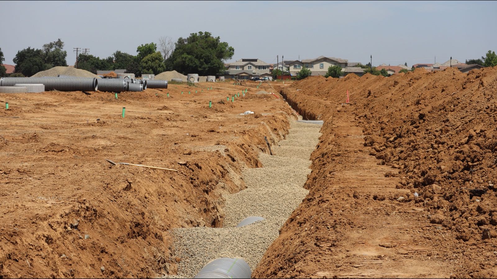 Long gravel-bedded trench with drain pipe cut through a graded red-dirt lot, pipe sections stacked behind
