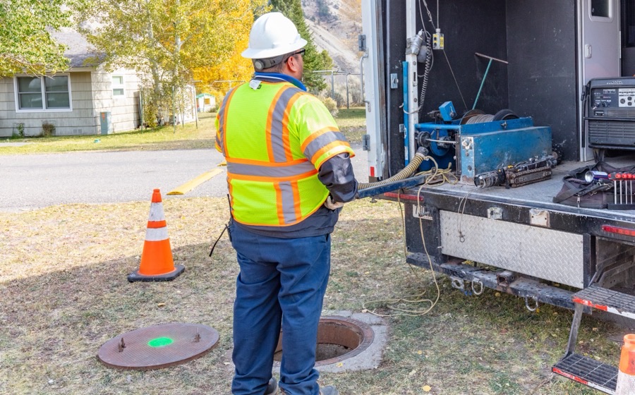 Worker in a hi-vis vest and hard hat beside a vacuum truck, an orange cone and an open manhole