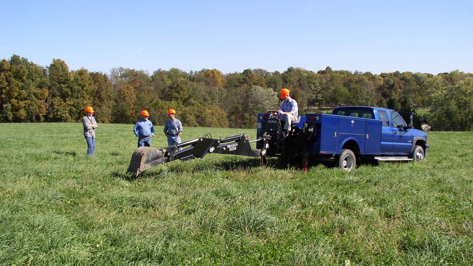 Four workers in orange hard hats on a grass field beside a blue service truck with a backhoe arm